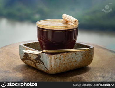 Espresso with milk foam served with Crispy Stick Candy on a Transparent Glass and Brass Spoon on Ceramic Saucer with Nature background. Coffee Time in the Morning, Space for text, Selective focus.