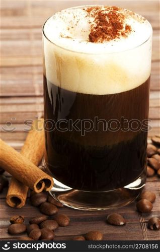 espresso with milk froth cocoa powder and cinnamon sticks. espresso in a straigt glass with milk froth cocoa powder, cinnamon sticks and coffee beans aside on wooden background