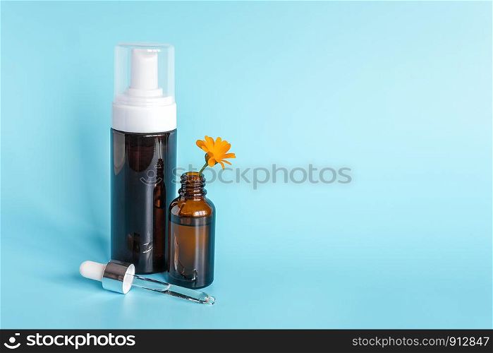 Essential oil in open brown dropper bottle with lying glass pipette, big bottle with white dispenser and orange flower calendula on blue background. Concept natural organic beauty cosmetics product.. Essential oil in open brown dropper bottle with lying glass pipette, big bottle with white dispenser and orange flower calendula on blue background. Concept natural organic beauty cosmetics product