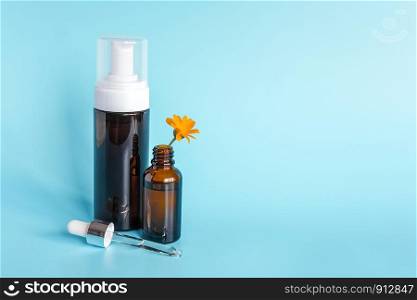 Essential oil in open brown dropper bottle with lying glass pipette, big bottle with white dispenser and orange flower calendula on blue background. Concept natural organic beauty cosmetics product.. Essential oil in open brown dropper bottle with lying glass pipette, big bottle with white dispenser and orange flower calendula on blue background. Concept natural organic beauty cosmetics product