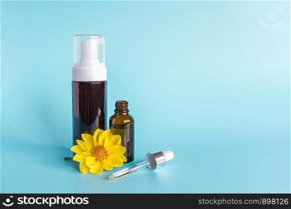 Essential oil in small open brown dropper bottle with lying glass pipette, big bottle with white dispenser and yellow flower on blue background. Concept natural organic beauty cosmetics product.. Essential oil in small open brown dropper bottle with lying glass pipette, big bottle with white dispenser and yellow flower on blue background. Concept natural organic beauty cosmetics product