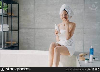 European beauty relaxes, laughs in bathroom. Attractive woman, towel-wrapped, applies lotion. Home shower, anti-cellulite massage, spa.