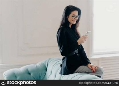 European entrepreneur in corporate suit and spectacles, using online banking app for money transfer, holding cellphone, sending emails, posing at the back of a comfortable sofa, utilizing free wifi.