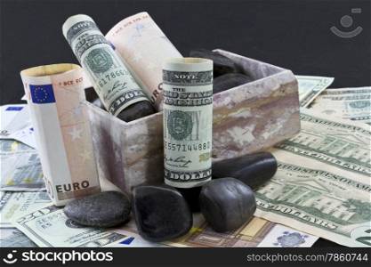 European euros and American dollars sit on a bed of currency within a marble box accented with black rocks suggesting difficult financial trends in international banking, investment, and business. Black background and upper area copy space.