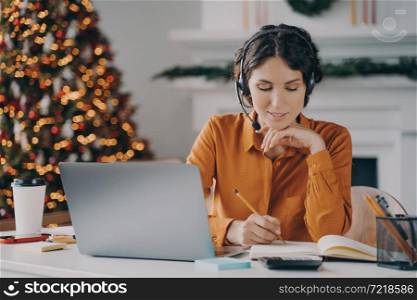 European female call center oparator in headset with mic making notes in agenda while speaking with client via laptop online, works from home with Xmas tree on background. Work during winter holidays. European female call center oparator in headset with mic working during winter holidays