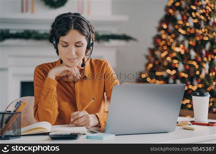 European female call center oparator in headset with mic making notes in agenda while speaking with client via laptop online, works from home with Xmas tree on background. Work during winter holidays. European female call center oparator in headset with mic working during winter holidays