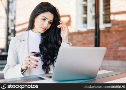 European female freelancer thinking on ideas for her new project working with laptop computer while sitting at outdoor cafeteria and drinking takeaway coffee having serious and thoughtful expression