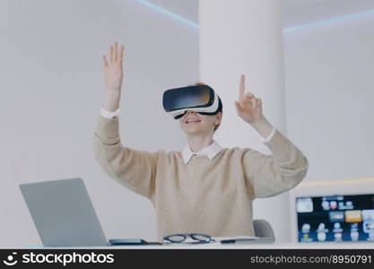 European girl in VR headset click buttons in virtual reality. Happy girl is sitting at the desk in front of laptop. Entrepreneur working on visual effects project. Concept of digital solutions.. European girl in VR headset click buttons in virtual reality. Working on visual effects project.