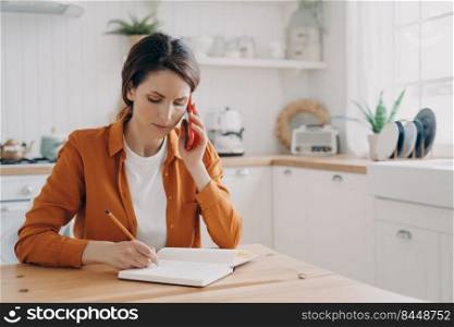 European girl is sitting at the kitchen, taking notes and talking on phone. Tired young woman is remote employee. Secretary, office manager or customer support assistant. Distance work concept.. Girl is sitting at the kitchen, taking notes and talking on phone. Office manager distance work.
