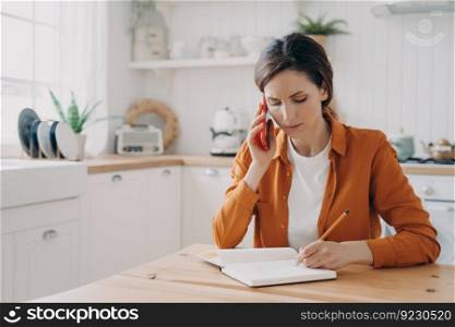 European girl is sitting at the kitchen, taking notes and talking on phone. Tired young woman is remote employee. Secretary, office manager or customer support assistant. Distance work concept.. Girl is sitting at the kitchen, taking notes and talking on phone. Office manager distance work.
