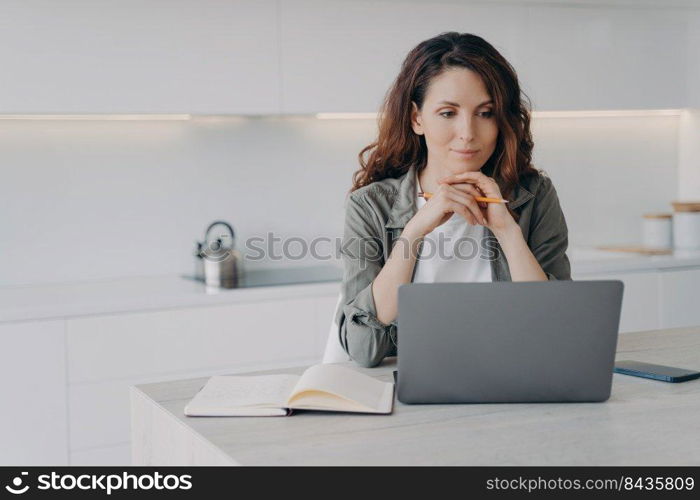 European girl is sitting in front of screen with hands folded. Confident female executive is working with laptop at home. Successful lady boss at her kitchen. Remote work of lawyer assistant.. European girl is sitting in front of screen with hands folded. Remote work of lawyer assistant.