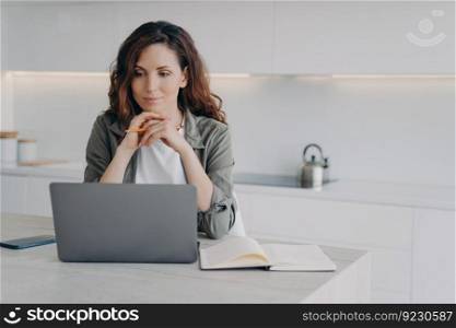 European girl is sitting in front of screen with hands folded. Confident female executive is working with laptop at home. Successful lady boss at her kitchen. Remote work of lawyer assistant.. European girl is sitting in front of screen with hands folded. Remote work of lawyer assistant.