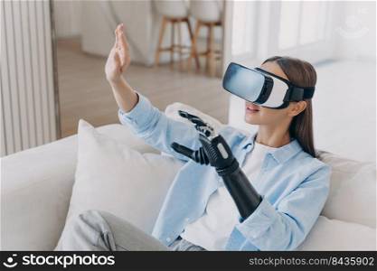 European handicapped girl in vr glasses on sofa at home. Disabled person gets rehabilitation for sensor cyber arm. Young woman is playing game. Healing technology, myoelectric robotic limb.. European handicapped girl in vr glasses on sofa at home. Healing technology, robotic limb.