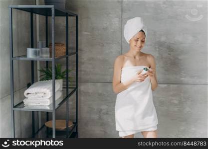 European lady applies cream holding a jar. Attractive caucasian girl wrapped in towel after bathing and hair washing. Happy young woman takes shower at home. Daily skincare and hygiene concept.. European lady applies cream. Happy young woman wrapped in towel after bathing and hair washing.