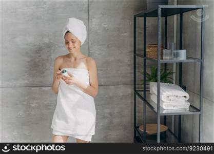 European lady applies cream holding a jar. Attractive caucasian girl wrapped in towel after bathing and hair washing. Happy young woman takes shower at home. Daily skincare and hygiene concept.. European lady applies cream. Happy young woman wrapped in towel after bathing and hair washing.