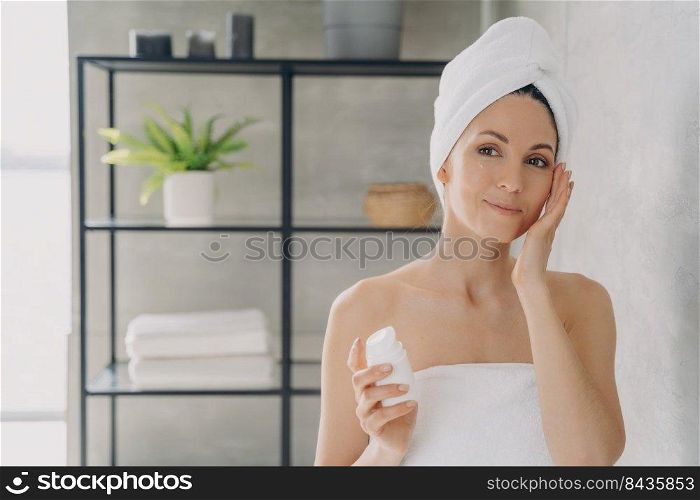 European lady applies face lotion or cream from bottle and looking into mirror. Face moisturizer, lifting cream. Attractive girl wrapped in towel after bathing and hair washing. Spa resort.. Face moisturizer, lifting cream. European lady applies face lotion or cream from bottle.