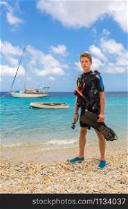 European male diver standing on beach of Bonaire with sea and boats