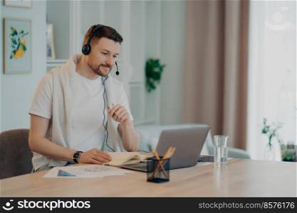 European male student learns foreign languages online has video lesson uses modern headset focused at laptop screen computer engaged in conference listens lecture attentively makes notes in diary