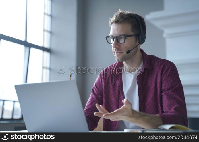 European man online tutor wearing glasses sitting at desk in headset and talking by video call on laptop computer while working remotely from home. Distance education and e-learning concept. European man online tutor wearing glasses sitting at desk in headset and talking by video call