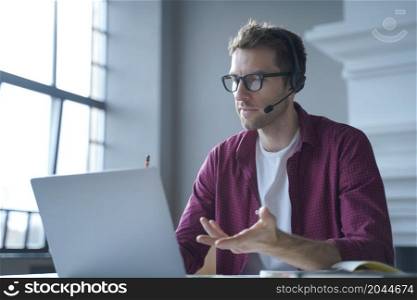 European man online tutor wearing glasses sitting at desk in headset and talking by video call on laptop computer while working remotely from home. Distance education and e-learning concept. European man online tutor wearing glasses sitting at desk in headset and talking by video call