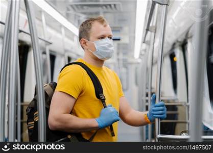 European man wears surgical mask, commutes in metro or underground, carries backpack, protects himself from viruses or epidemic disease, stands in empty carriage, follows social distancing.