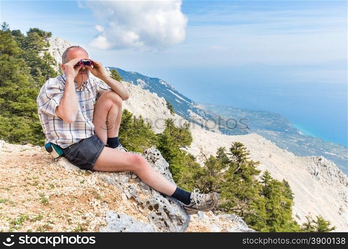 European middle aged man sitting on rock in mountains looking at landscape through binoculars