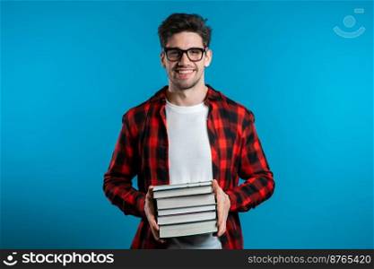 European student in red plaid shirt on blue background in studio holds stack of university books from library. Guy smiles, he is happy to graduate. European student in red plaid shirt on blue background holds stack books