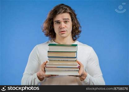 European student is dissatisfied with amount of homework and books on blue background. Man with long hairdo in displeasure, he is annoyed,discouraged frustrated by studies. High quality photo. European student is dissatisfied with amount of homework and books on blue background. Man with long hairdo in displeasure, he is annoyed,discouraged frustrated by studies