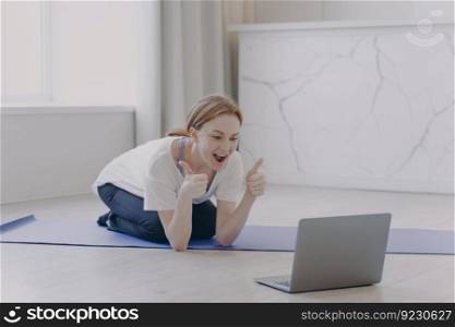 European white woman has distance sport classes. Girl gestures thumbs and feels good. Internet yoga lesson on mat. Concept of e-learning motivation. Sport online while coronavirus quarantine.. European white woman has distance sport classes. Girl gestures thumbs and feels good.