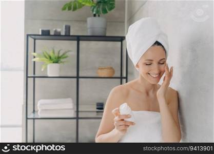 European woman applies face lotion, attractive girl with towel after bathing, happy lady showers at home. Daily skin care.