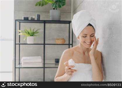 European woman applies face lotion or cream from flacon and smiling. Attractive caucasian girl wrapped in towel after bathing and hair washing. Happy young lady takes shower at home. Daily skin care.. European woman applies face lotion or cream from flacon and smiling. Daily skin care.