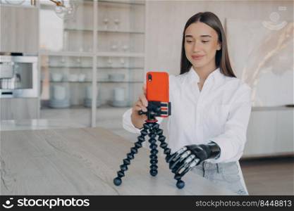 European woman is filming with phone on tripod. Attractive handicapped girl. Amputee is taking video how to use cyber arm. Beauty blogger has modern bionic prosthesis. Equality concept.. European woman is filming with phone. Attractive handicapped girl has modern bionic prosthesis.