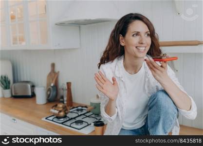 European young woman talking on telephone. Happy girl with telephone sitting on worktop of table at kitchen and having conversation. Modern furniture and cosy interior design. Lifestyle concept.. Happy european girl with telephone sitting on worktop of table at kitchen and having conversation.