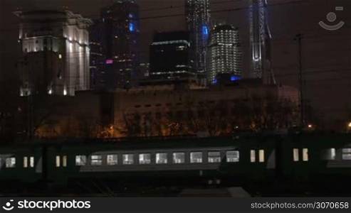 Evening commuter train and railroad engine passing by in metropolis with modern illuminated skyscrapers