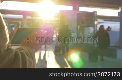 Evening time at the station. Woman typing message on touch pad, people walking around and bright sun flare in the sky