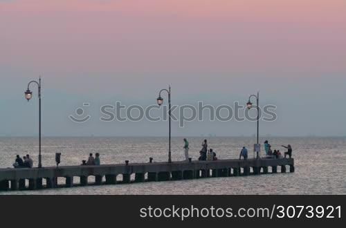 Evening time on the coast. People relaxing on the stone pier with lit lanterns and looking at quiet sea