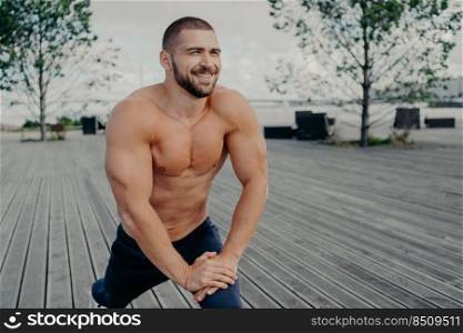 Everyday morning routine and sport exercises concept. Sporty unshaven man bodybuilder with cheerful expression does stretching exercises for legs, poses with naked torso, has workout outdoor.