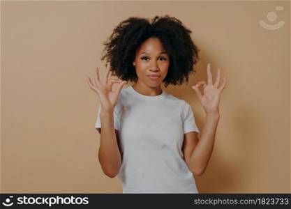 Everything is fine. Young confident african american lady in white tshirt keeping both hands in okay gesture, making ok sign while standing isolated over brown background in studio. Young african american lady keeping both hands in okay gesture, isolated over brown background