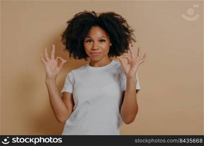 Everything is fine. Young confident african american lady in white tshirt keeping both hands in okay gesture, making ok sign while standing isolated over brown background in studio. Young african american lady keeping both hands in okay gesture, isolated over brown background