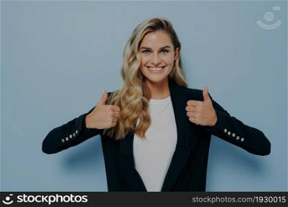 Excellent job gesture. Cheerful girl with blonde wavy hair looking at camera with satisfaction, smiling and showing thumbs up with both hands. Body language concept and positiveness. Cheerful girl with blonde hair looking at camera with satisfaction, smiling and showing thumbs up