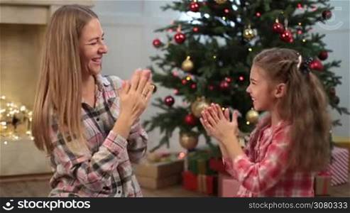 Excited adorable girl with brown hair playing patty cake with her attractive smiling mother over Christmas decorated room background. Mom having fun and playing pat-a-cake with her cute little daughter during winter holidays at home. Dolly shot.