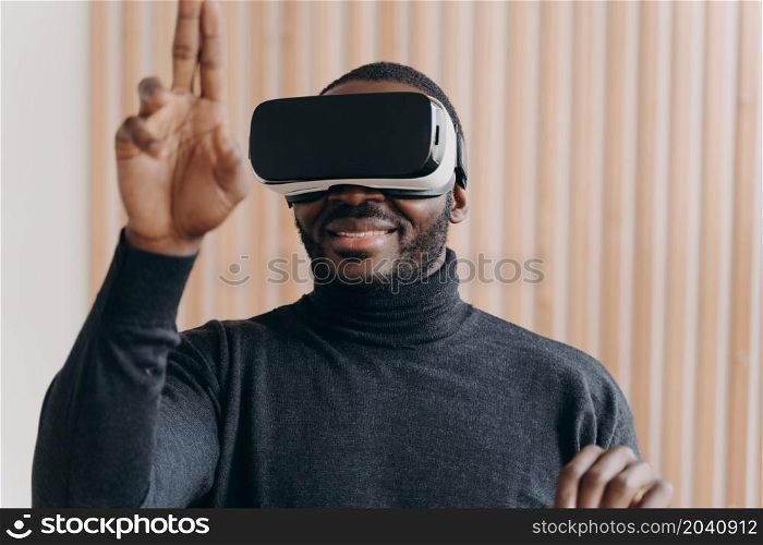 Excited African American man in VR headset glasses enjoying augmented reality while sitting at modern workplace at home, cheerfully raised hand with two fingers up as interacting with cyberspace in 3D. Excited African American man in VR headset glasses enjoying augmented reality in office
