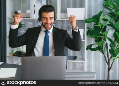 Excited and happy businessman dressed in black formal suit, raise his arm in celebratory gesture at office desk. Successfully call center or telesales operator celebrate in workplace. Fervent. Excited and happy businessman dressed in black formal suit. Fervent