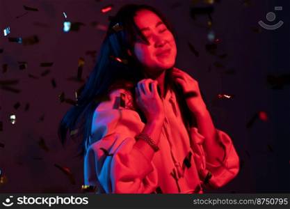 Excited asian attractive woman dancing, having fun, rejoices over confetti rain with neon light in studio. Concept of happiness, party, winning.. Excited asian attractive woman dancing, having fun, rejoices over confetti rain with neon light in studio. Concept of happiness, party, winning