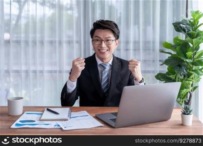 Excited Asian businessman celebrates success at office desk. Happy office worker achieves business goals with data analysis or marketing planning. Business winner celebratory expression. Jubilant. Excited Asian businessman celebrates success at office desk. Jubilant