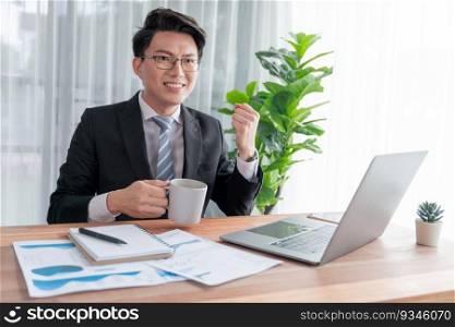 Excited Asian businessman celebrates success at office desk. Happy office worker achieves business goals with data analysis or marketing planning. Business winner celebratory expression. Jubilant. Excited Asian businessman celebrates success at office desk. Jubilant