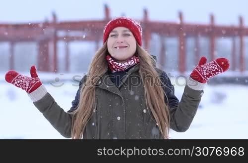 Excited attractive blonde woman standing with opened arms outdoors and catching snowflakes with hands. Cheerful female enjoying first snow and watching snowflakes falling on snowy winter day.