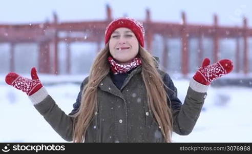 Excited attractive blonde woman standing with opened arms outdoors and catching snowflakes with hands. Cheerful female enjoying first snow and watching snowflakes falling on snowy winter day.