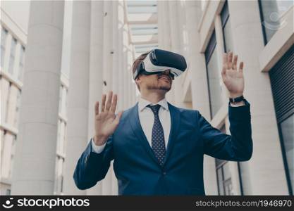 Excited business person in formal clothes walking around in virtual reality while wearing VR goggles, holding both of his hands in air, interacting with digital world, standing alone between buildings. Man in formal blue suit walking around in virtual reality while wearing VR goggles