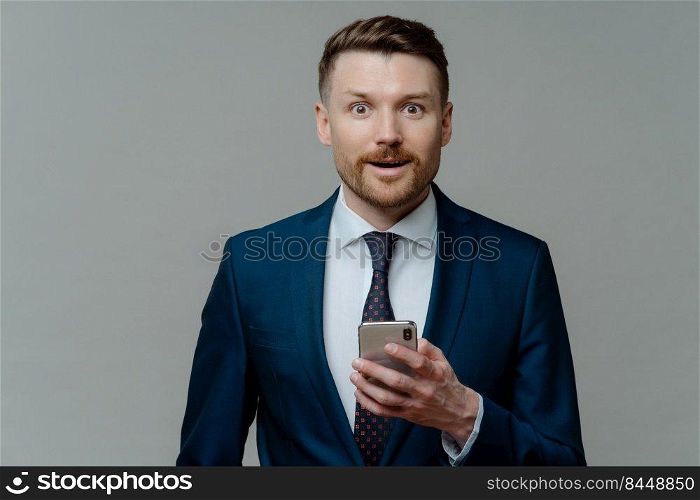 Excited businessman in elegant clothes using smartphone, looking at camera with surprised face expression, male ceo executive reading good news on mobile phone while standing against grey background. Amazed businessman in suit reading good news on smartphone